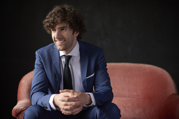 Studio shot of a confident young businessman wearing suit while sitting on sofa at black background.