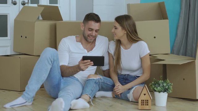 Young happy couple sitting on floor using tablet computer to find transport service for reloction in their new home