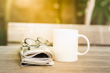 Cup of hot coffee and newspaper and reading glasses on wooden table in garden morning