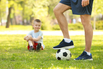 Young man with soccer ball in green park and his blurred son on background