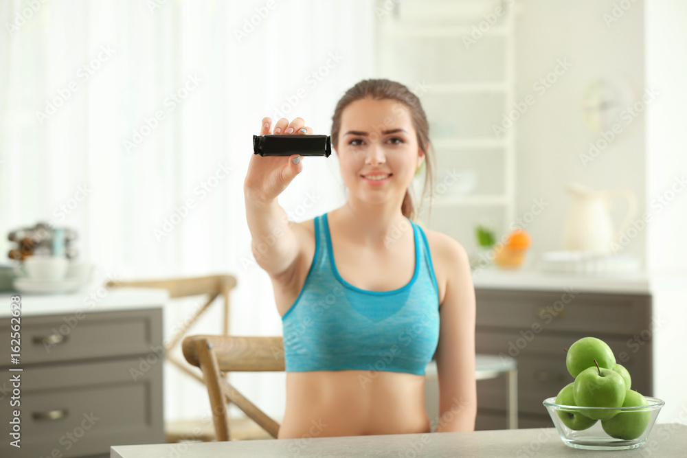 Canvas Prints Beautiful young woman with energy bar sitting at table in kitchen. Diet concept