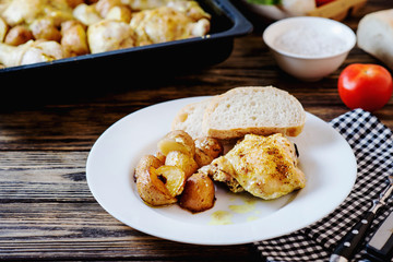Hearty and delicious dinner, baked chicken legs and thighs with potatoes, onions, tomatoes, spices, herbs and white bread on a dark wooden background
