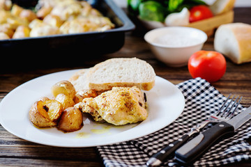 Hearty and delicious dinner, baked chicken legs and thighs with potatoes, onions, tomatoes, spices, herbs and white bread on a dark wooden background