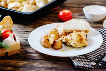 Hearty and delicious dinner, baked chicken legs and thighs with potatoes, onions, tomatoes, spices, herbs and white bread on a dark wooden background