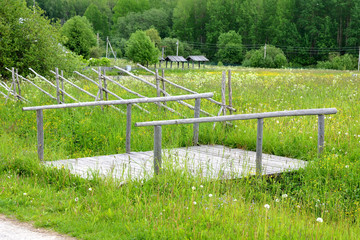 Wooden bridge of planks and thin poles across a ditch next to the meadow
