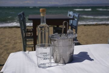 Ouzo, tsipouro bottle with empty glass and ice cubes on tavern table by the sea.
Glass bottle of ouzo, tsipouro drink by Greek beach with blurred tavern table and wooden blue chairs background.