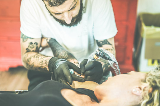 Bearded tattoo artist making tattoo inside ink studio on young blond woman - Tattooer at work - Skin trend fashion concept - Focus on man hands machine - Vintage cine filter