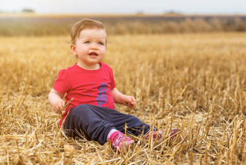 Naklejka na ściany i meble Happy baby girl sitting on harvested wheat field with sun in the background