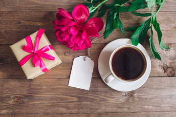 A cup of coffee , pink peonies pattern and gift box on wooden background . Good morning. Women's or Mother's Day background. Top view