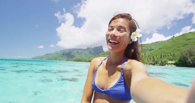 Selfie tropical vacation bikini woman smiling at camera at luxury travel resort in tropical summer. Happy girl relaxing with polynesian flower, Asian beauty. RED EPIC SLOW MOTION.