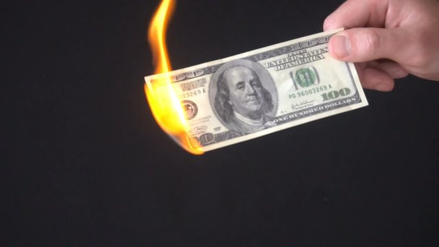Hand holding 100 dollars on a black background and sets it on fire with a lighter. Hand holding burning one hundred dollars.