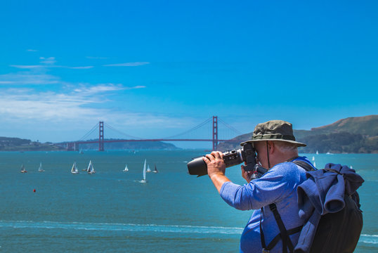 A male photography using a camera with a long lens takes pictures on Angel Island across the San Francisco Bay from the landmark Golden Gate Bridge