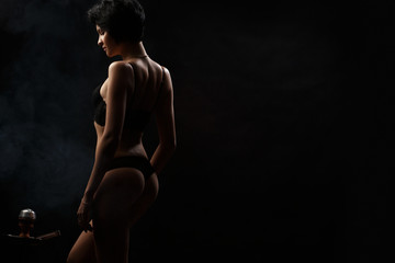 Fototapeta premium Sexy, beautiful and attractive woman with gorgeous slim body wearing lacy underwear is posing in the dark studio. Appealing female model with seductive figure and short haircut in the black lingerie