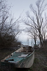 An old, damaged  and abandoned fishing boat,  near a lake shore, with some trees and others boats nearby