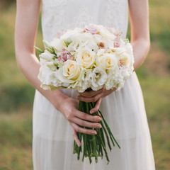 bride in a dress standing in a green garden and holding a wedding bouquet of flowers and greenery....