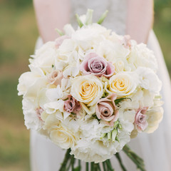 bride in a dress standing in a green garden and holding a wedding bouquet of flowers and greenery. Woman holding colorful bouquet with her hands on wedding day