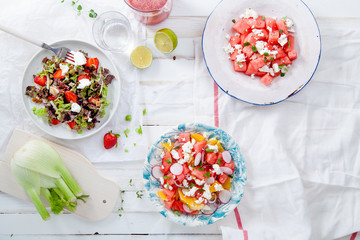 Variety of raw fresh summer salads with fruits and berries on white wooden background served with napkins and drinks. Summertime organic easy lifestyle concept. Flat lay with copy space.