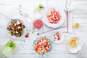 Variety of raw fresh summer salads with fruits and berries on white wooden background served with napkins and drinks. Summertime organic easy lifestyle concept. Flat lay with copy space.