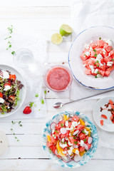 Variety of raw fresh summer salads with fruits and berries on white wooden background served with napkins and drinks. Summertime organic easy lifestyle concept. Flat lay with copy space.