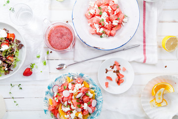 Variety of raw fresh summer salads with fruits and berries on white wooden background served with napkins and drinks. Summertime organic easy lifestyle concept. Flat lay with copy space.