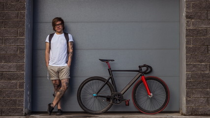 Tattooed biker hipster man in shorts standing next to a fixed gear bike against the background of gray rolling gates