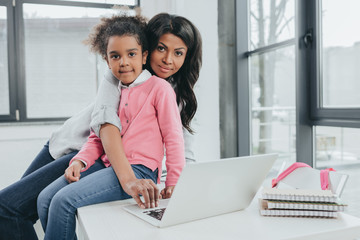 african american mother and daughter using laptop and looking at camera