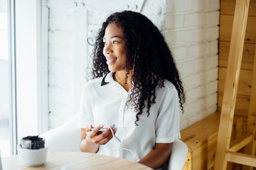 Young gorgeous african american woman with headphones looking in window sitting in modern loft interior of cafe and waiting for order.