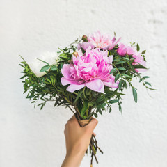 Bouquet of pink and white peony flowers in woman's hand, white wall background, copy space, square crop. Flower greeting card concept
