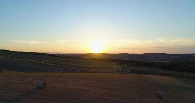Forward aerial view of green wheat rural fields hills with hay bales and sun. Nature outdoors travel destination Val d'Orcia, Tuscany, Italy. sunset or sunrise sunny summer or spring. 4k drone video