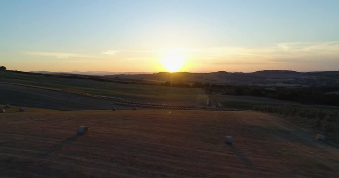 Backward aerial view of green wheat rural fields hills with hay bales and sun. Nature outdoors travel destination Val d'Orcia, Tuscany, Italy. sunset or sunrise sunny summer or spring. 4k drone video