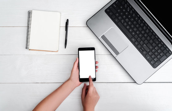 Top view on white wooden table with open blank laptop computer, cell phone, empty diary and girl's hands with pen, free space. Girl's hands writing in diary. Mobile phone with white screen copy space