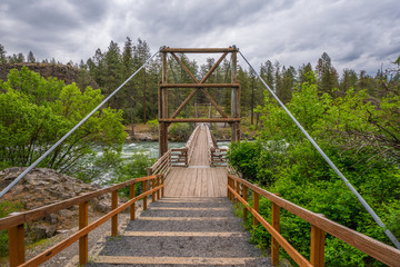 Wooden bridge across the river on a background of the green forest. Dark blue rough river. Scenic landscape. Riverside state park, Bowl and Pitcher, Spokane area, Eastern Washington, USA.