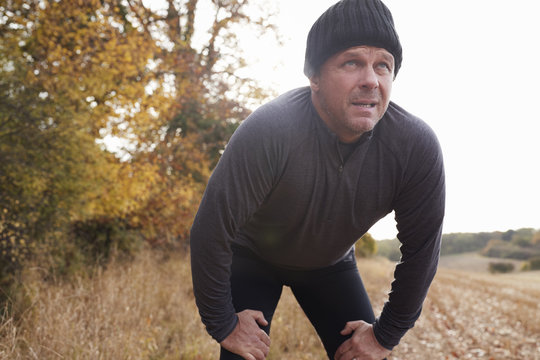 Mature Male Runner Pausing For Breath During Exercise In Woods