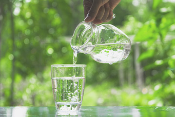 Female hand pouring water from pitcher to glass on nature background - obrazy, fototapety, plakaty