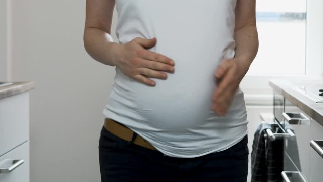 A pregnant woman wearing a white t-shirt and black pants dances in her kitchen. A close up shot of her body as she sways her hips and taps her body.