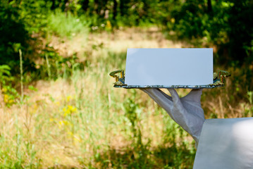 A waiter's hand in a white glove and with a white napkin holds a metal tray of silvery rectangular metal with a white sign on a blurred background of nature green bushes and trees