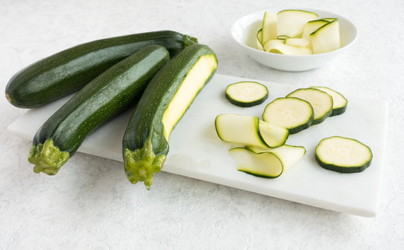 Whole zucchini, curls and slices on white marble chopping board and white marbled background. 