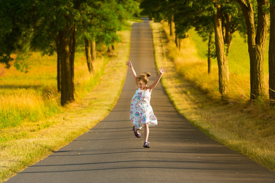 Kleines M&auml;dchen in Sommerkleid l&auml;uft springend und jubelnd eine Stra&szlig;e entlang und freut sich an einem warme Sommerabend - Pure Lebensfreude!