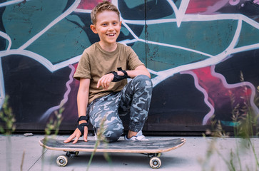 Smiling boy with skateboard sits near grafity painted wall
