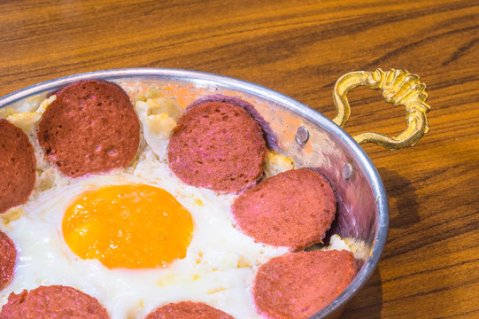 Closeup of Turkish sausage (sucuk) and fried egg in a copper pan on wooden table