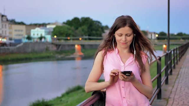 Pretty young girl using smartphone with earphones near the railing on waterfront in evening time, old city, river and bridge in the background