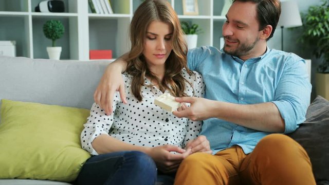 Attractive man surprising his emotional girlfriend with a gift while they relaxing on sofa at home in the living room