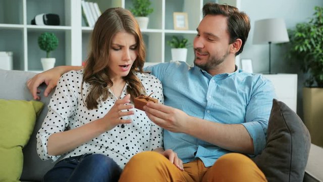 Young man surprising his emotional girlfriend with a gift, happy woman hugging and kissing her boyfriend while they relaxing on sofa at home.