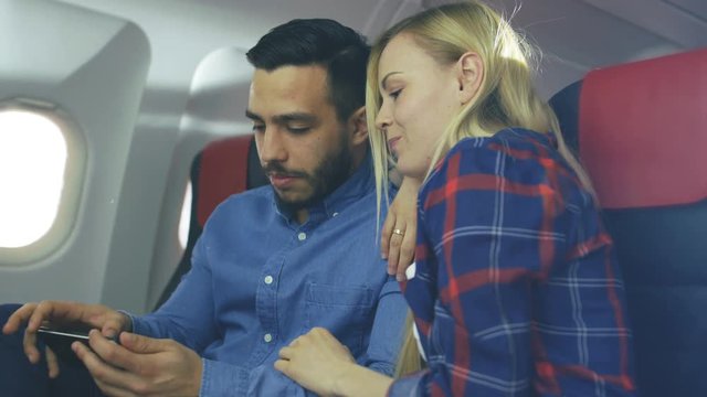 On a Board of Commercial Airplane Beautiful Young Blonde with Handsome Hispanic Male Play Games on Smartphone and Smile. Sun Shines Through Aeroplane Window. 4K UHD.