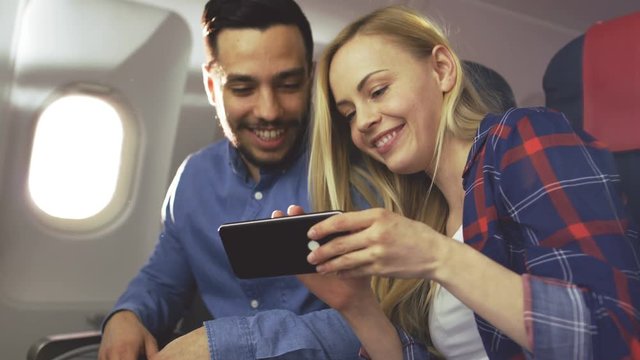 On a Board of Commercial Airplane Beautiful Young Blonde with Handsome Hispanic Male Share Videos on Smartphone and Smile. Sun Shines Through Aeroplane Window. 4K UHD.