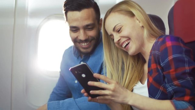 On a Board of Commercial Airplane Beautiful Young Blonde Shows Her Smartphone Screen to a  Handsome Hispanic Male they Both Laugh. Sun Shines Through Aeroplane Window. 4K UHD.