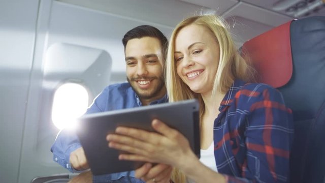 On a Board of Commercial Airplane Beautiful Young Blonde with Handsome Hispanic Male Use Tablet Computer and Smile. Sun Shines Through Aeroplane Window. Shot on RED EPIC-W 8K Helium Cinema Camera.