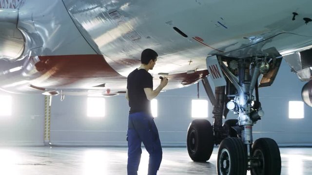 In a Hangar Aircraft Maintenance Engineer/ Technician/ Mechanic Visually Inspects Airplane's Chassis. Shot on RED EPIC-W 8K Helium Cinema Camera.