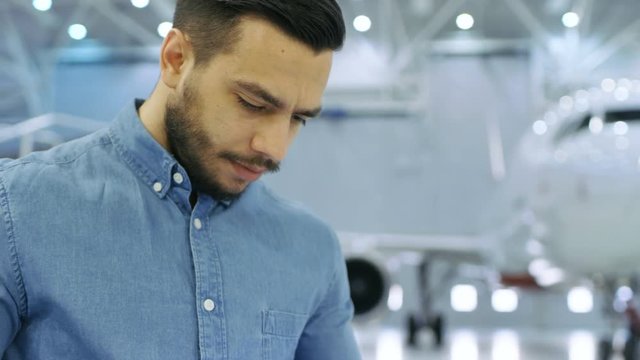 In Big Company Hangar Aircraft Maintenance Engineer Uses Tablet Computer while Standing Near Big New Shiny White Plane. Shot on RED EPIC-W 8K Helium Cinema Camera.