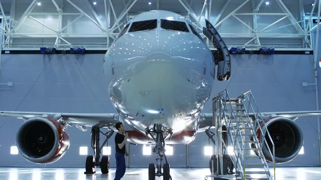 Up-Down Shot of a Brand New Airplane Standing in a Aircraft Maintenance Hangar while Aircraft Maintenance Engineer/ Technician/ Mechanic Visually Inspects Plane with a Flashlight. 